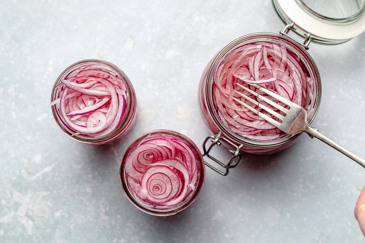 An overhead shot of 3 jars or easy pickled red onions on a light blue surface. One jar is larger than the other two and there is a fork pressing down on the red onions to help fully submerge the onions in the pickling liquid.