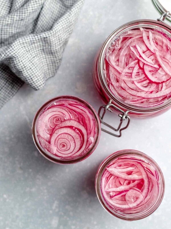 An overhead shot of three open jars of quick pickled red onions on a light blue surface. There is a light blue linen napkin in the upper left hand corner.