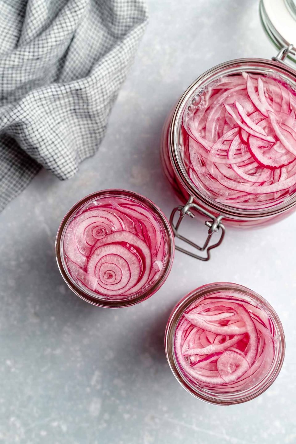 An overhead shot of three open jars of quick pickled red onions on a light blue surface. There is a light blue linen napkin in the upper left hand corner.