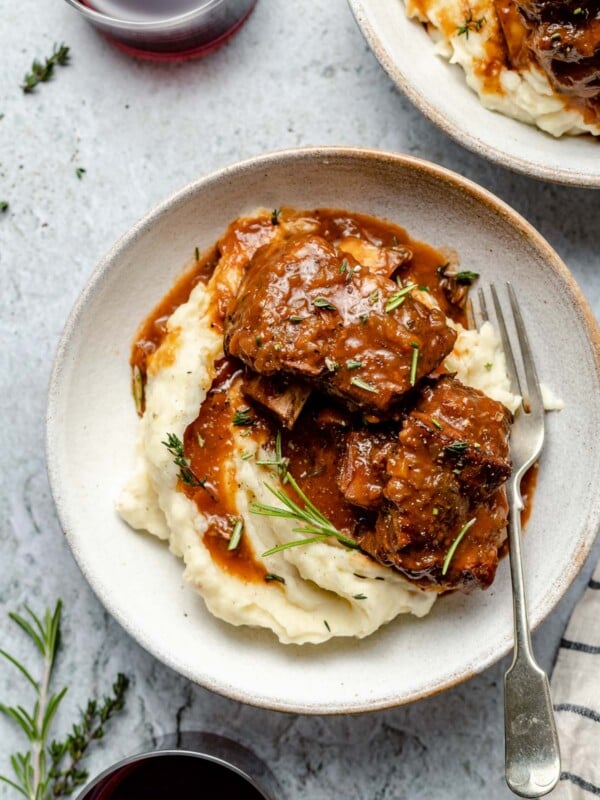 Two servings of Red Wine Braised Short Ribs atop a pile of mashed potatoes on two white ceramic plates. The plate at center has a single silver fork resting on top and both plates have been garnished with fresh herbs. A white and blue striped linen napkin, two glasses of red wine, and loose fresh herbs surround the plates and all items sit atop a blue-gray textured surface.