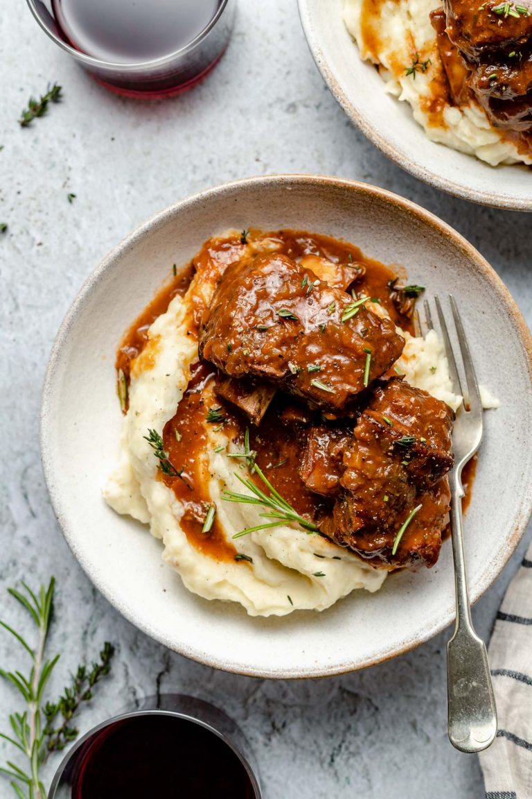 Two servings of Red Wine Braised Short Ribs atop a pile of mashed potatoes on two white ceramic plates. The plate at center has a single silver fork resting on top and both plates have been garnished with fresh herbs. A white and blue striped linen napkin, two glasses of red wine, and loose fresh herbs surround the plates and all items sit atop a blue-gray textured surface.