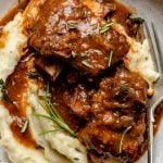 An overhead and close up shot of Red Wine Braised Short Ribs atop a pile of mashed potatoes served on a white ceramic plate. A single silver fork rests on top of the plate with fresh herbs sprinkled over top. The plate sits atop a blue-gray textured surface.
