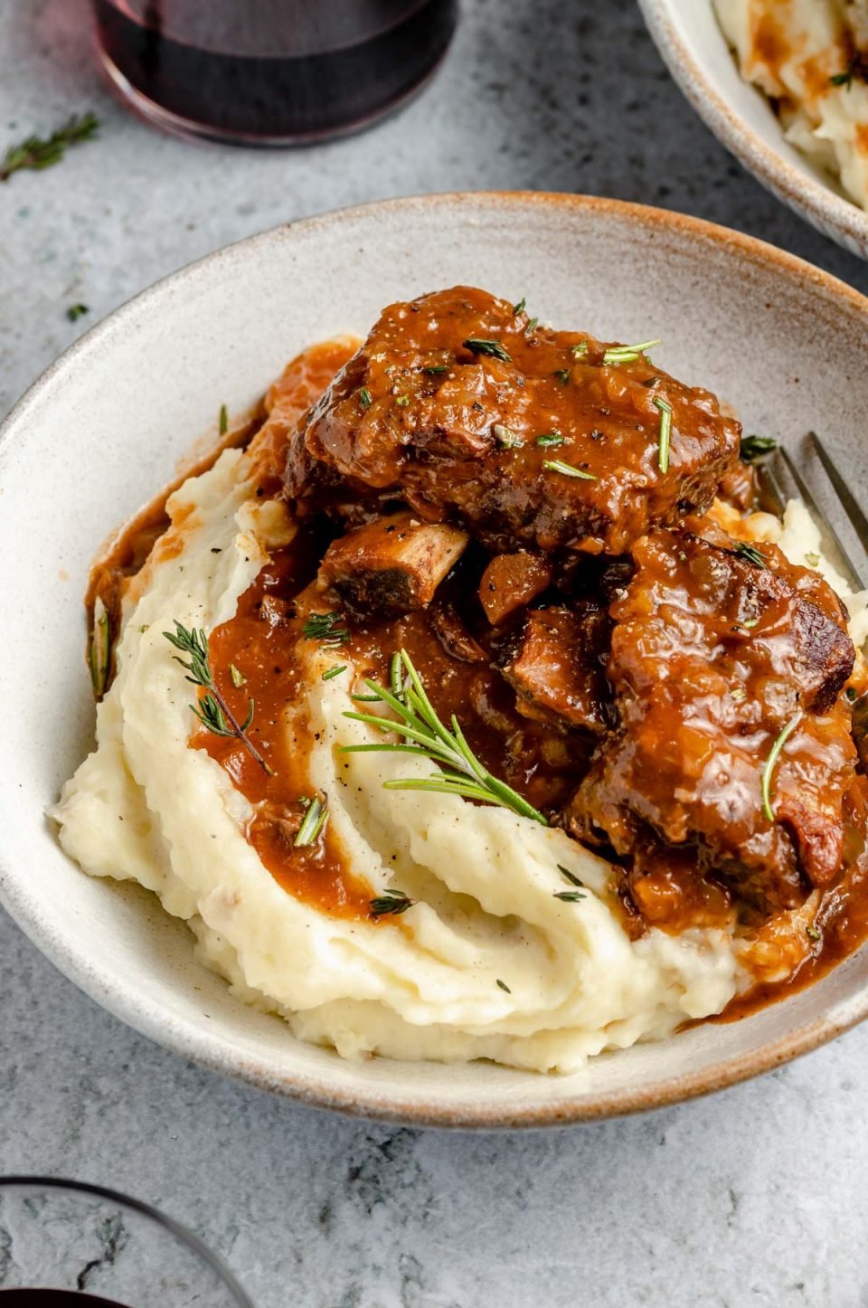 A side angle shot of Red Wine Braised Short Ribs atop a pile of mashed potatoes served in a creamy white ceramic bowl. A single silver fork rests inside the bowl with fresh herbs sprinkled over top. The bowl sits atop a blue-gray textured surface. Glasses of wine, another bowl of short ribs and loose fresh herbs surround the bowl at center.