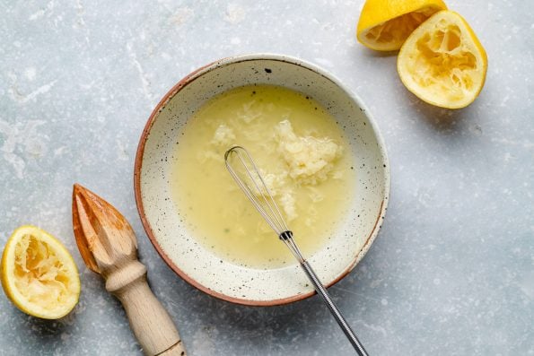 How to Make Go-To Lemon Tahini Sauce, step 1: Lemon juice & garlic are mixed in a ceramic bowl with a small mini whisk that rests inside of the bowl. Three lemon halves and a wooden citrus reamer surround the bowl.