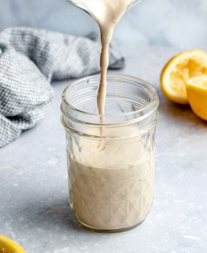 Lemon Tahini Sauce is poured into a clear glass Mason jar. The jar sits atop a light blue textured surface with a blue and white plaid linen napkin and three lemon halves surround the jar.