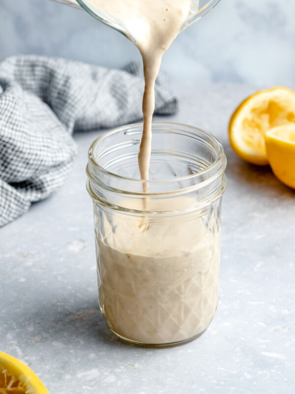 Lemon Tahini Sauce is poured into a clear glass Mason jar. The jar sits atop a light blue textured surface with a blue and white plaid linen napkin and three lemon halves surround the jar.