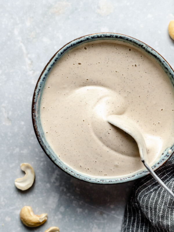 A blue speckled ceramic bowl rests on a light blue background and is filled with Vegan Cashew Crema. Loose raw cashews surround the bowl and a blue and white striped linen napkin is tucked against the bowl in the lower right hand corner. A spoon is being dipped into the bowl of vegan crema.