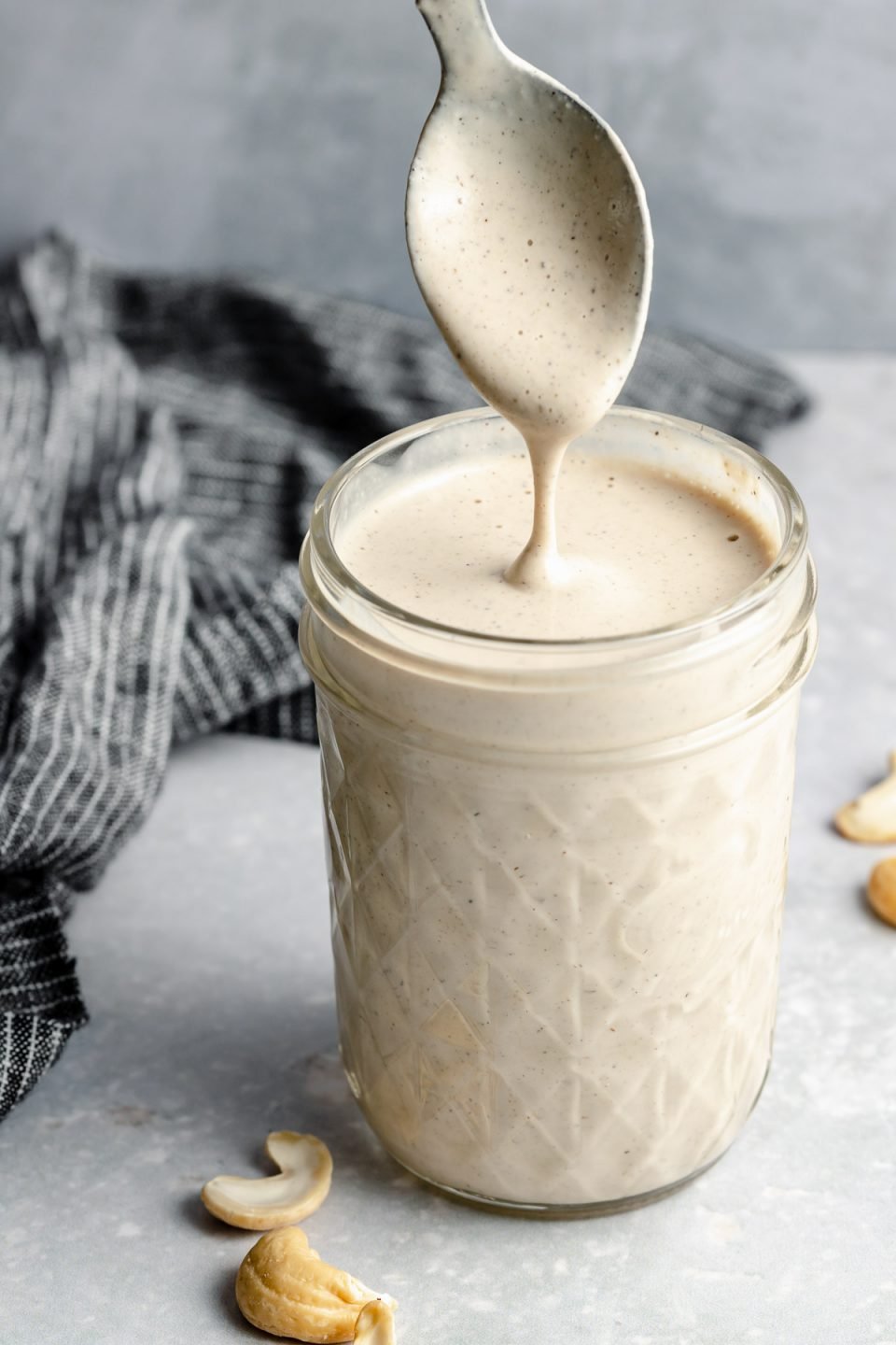 An angled shot of an open mason jar filled with Vegan Cashew Crema sits on top of a light blue surface. A spoon has been dipped into the crema and is being pulled out covered in the tan-colored creamy sauce & falling off the spoon back into the mason jar. A blue & white striped linen napkin rests on the surface in the background & raw cashews surround the jar of crema.