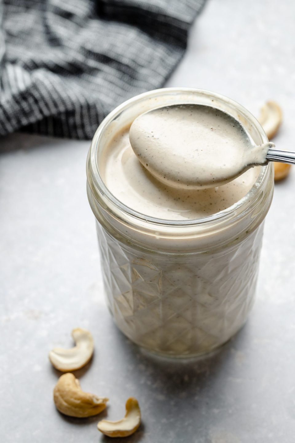 An overhead and angled shot of an open mason jar filled with Vegan Cashew Crema sits on top of a light blue surface. A spoon has been dipped into the vegan crema and a spoonful is hovering over the mason jar. A blue & white striped linen napkin rests on the surface in the background & raw cashews surround the jar of crema.