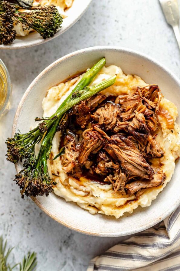 Two servings of shredded balsamic lamb are served over mashed potatoes in white ceramic bowls with broccolini and extra braising liquid over top. The bowls sit atop a gray textured surface with loose sprigs of rosemary, a silver fork, a striped linen napkin and a glass of white wine surrounds the bowls.