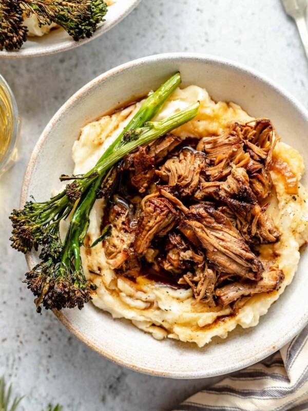 Two servings of slow cooked lamb are served over mashed potatoes in white ceramic bowls with broccolini and extra braising liquid over top. The bowls sit atop a gray textured surface with loose sprigs of rosemary, a silver fork, a striped linen napkin and a glass of white wine surrounds the bowls.