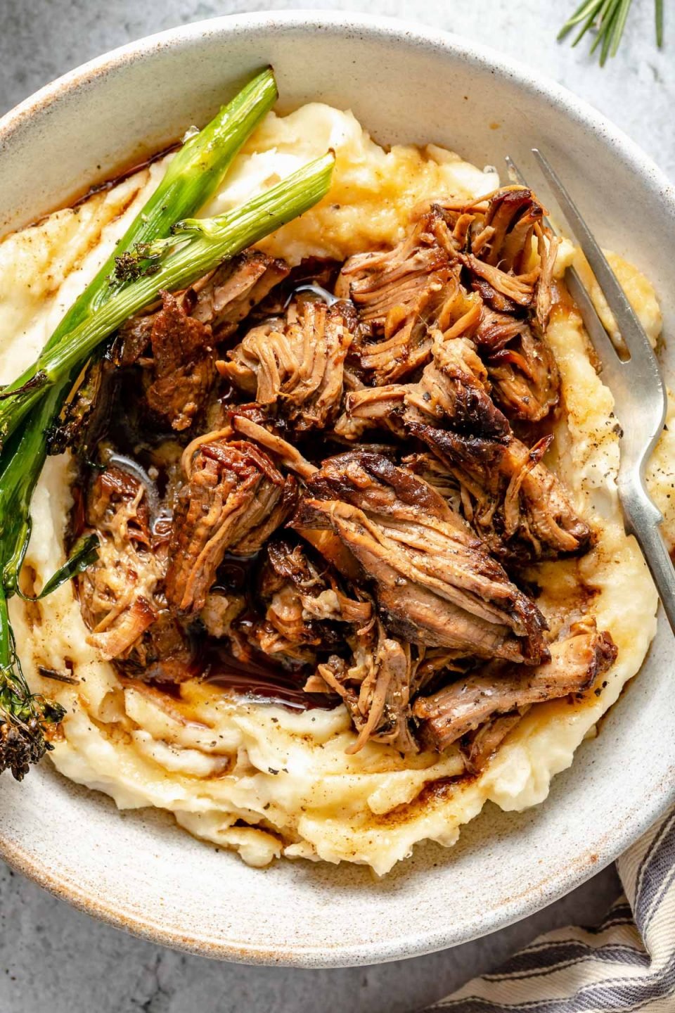A close up shot of finished balsamic lamb shoulder served over mashed potatoes in a white ceramic bowl with broccolini and extra braising liquid over top. The bowl sits atop a gray textured surface with loose sprigs of rosemary and a striped linen napkin surrounding the bowl. A silver fork rests inside of the bowl.