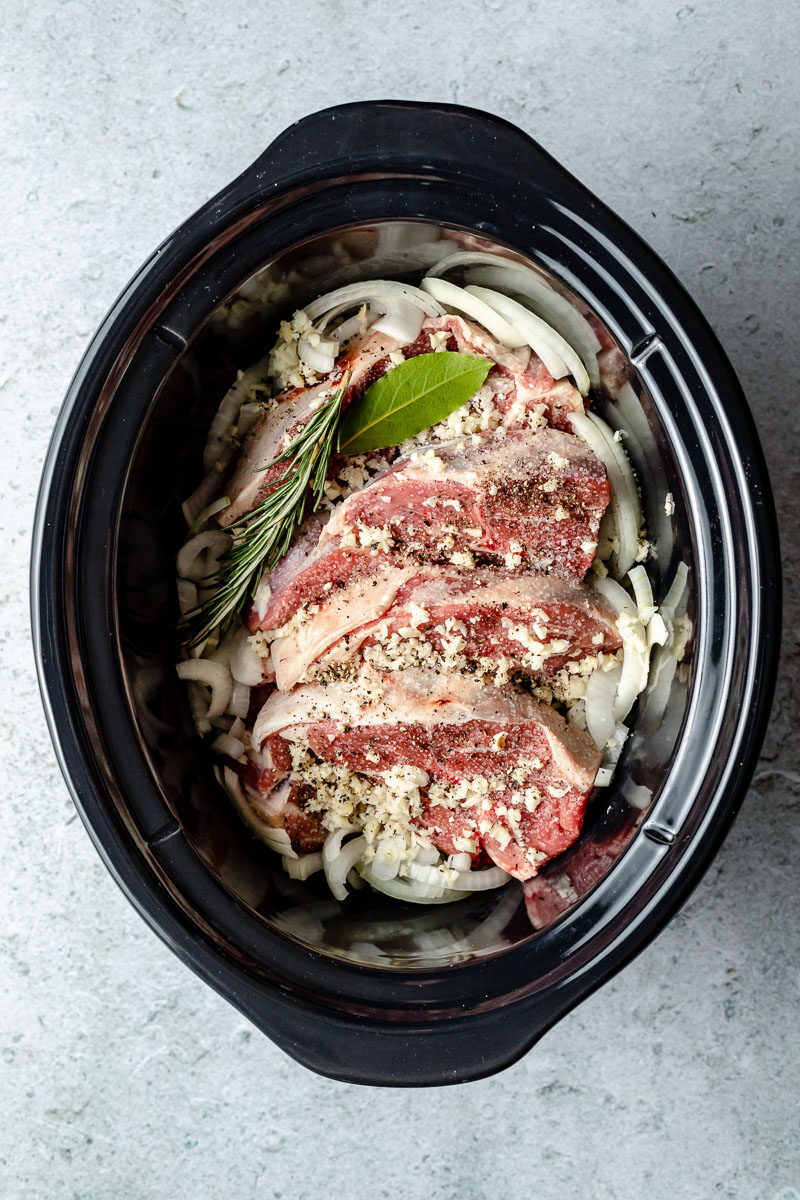 How to make crock pot lamb with balsamic, rosemary, and garlic, step 1: Assemble the lamb. Lamb shoulder, sliced onion, garlic, rosemary, bay leaf, kosher salt, and ground black pepper are placed inside a slow cooker.
