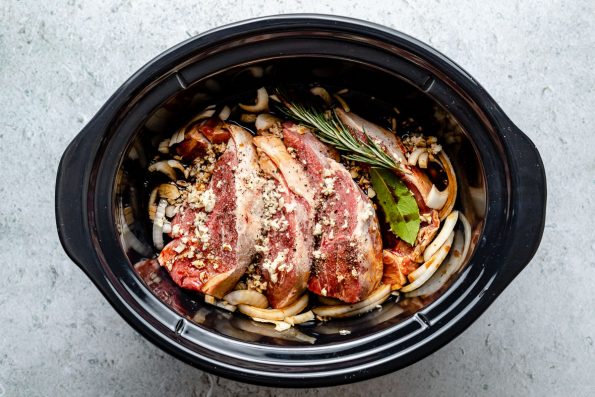 How to make Slow cooked lamb shoulder with balsamic, rosemary, and garlic, step 1: Assemble the lamb. Lamb shoulder, sliced onion, garlic, rosemary, bay leaf, kosher salt, and ground black pepper are placed inside a slow cooker with a braising liquid poured over.