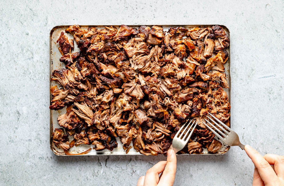 How to make Slow cooked lamb shoulder with balsamic, rosemary, and garlic, step 3: Shred the lamb. Shredded slow cooked lamb fills an aluminum baking sheet. A woman's hands hold two forks and uses them to shred the lamb. The baking sheet sits atop a grey textured surface.