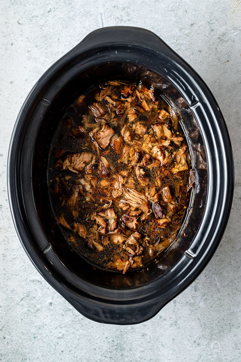 How to make Slow cooked lamb shoulder with balsamic, rosemary, and garlic, step 3: Shred the lamb. Shredded lamb fills a slow cooker in braising liquid. The slow cooker sits atop a grey textured surface.