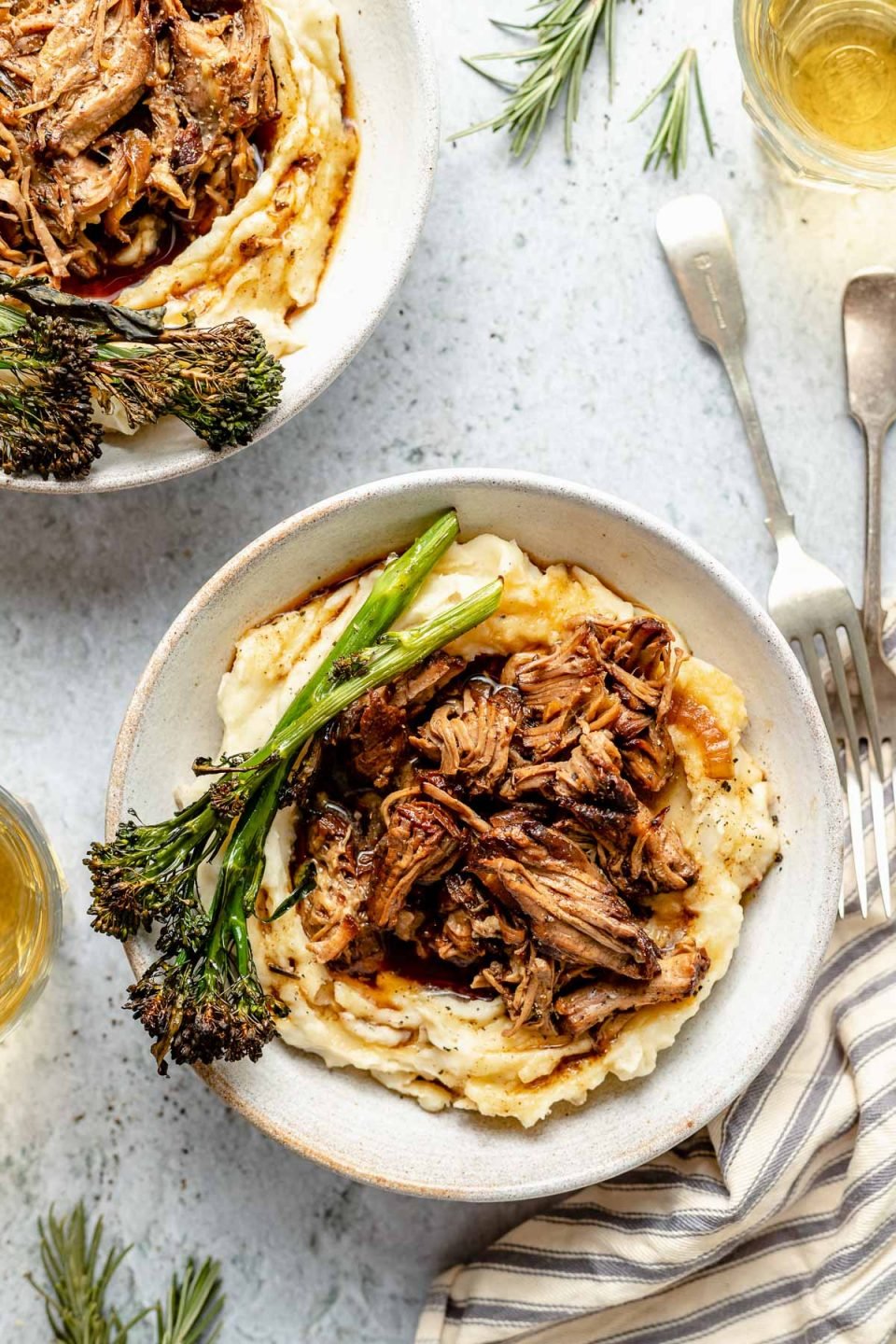 Two servings of slow cooked lamb are served over mashed potatoes in white ceramic bowls with broccolini and extra braising liquid over top. The bowls sit atop a gray textured surface with loose sprigs of rosemary, a silver fork, a striped linen napkin and a glass of white wine surrounds the bowls.