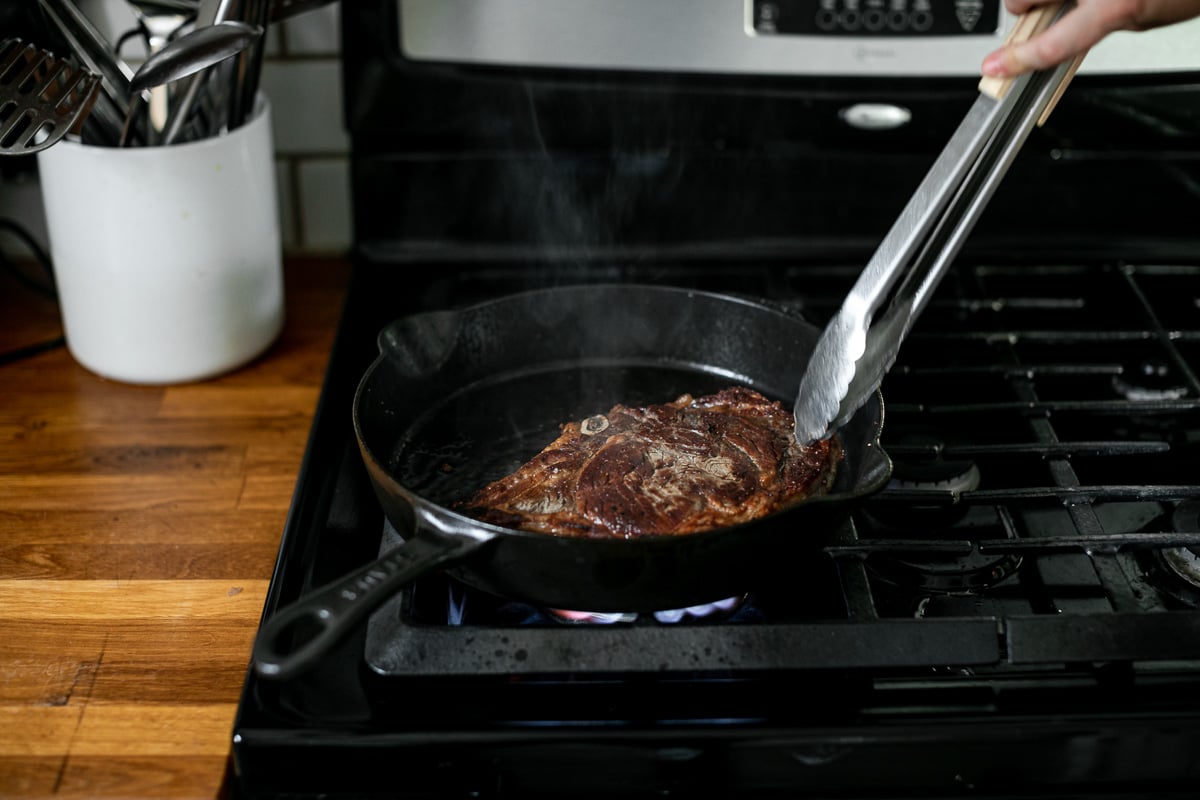 A woman's hand holds a pair of tongs, turning a seared steak in a cast iron skillet over high heat on a gas range.