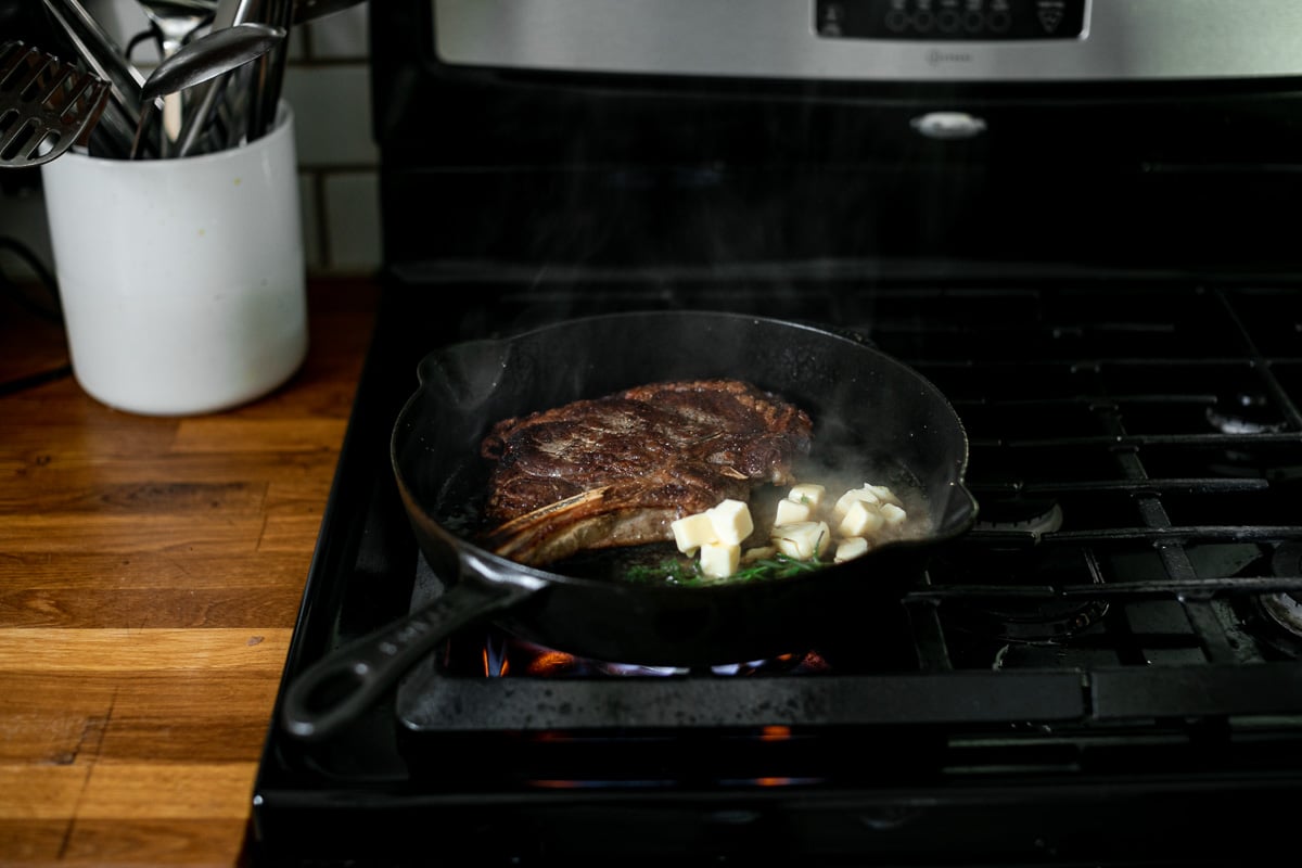A photo of a seared steak with butter and fresh herbs in a cast iron skillet over high heat on a gas range.