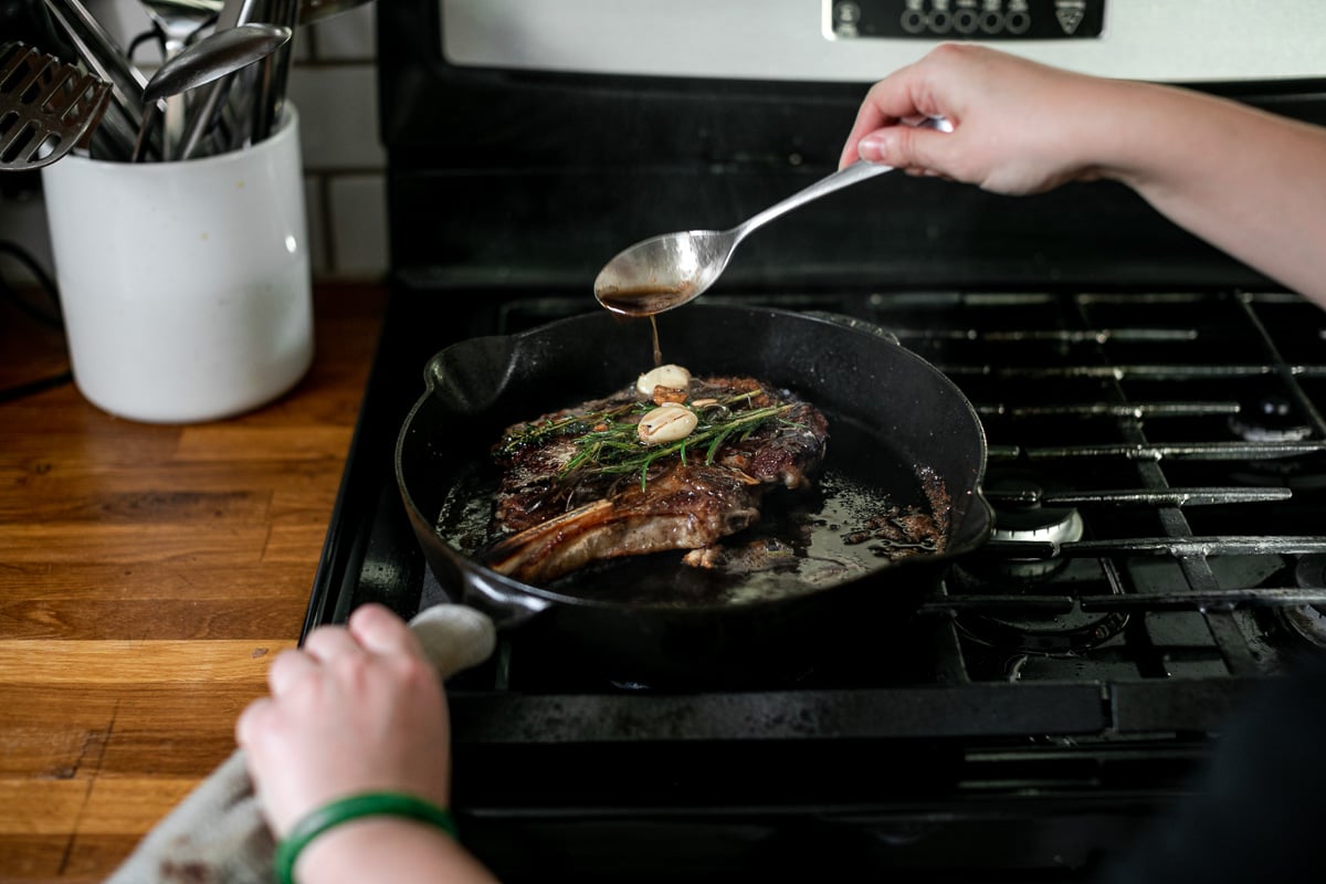 A woman's hands hold handle of skillet and spoon, spooning browned butter over the steak, which is topped with fresh herbs and crushed garlic cloves.