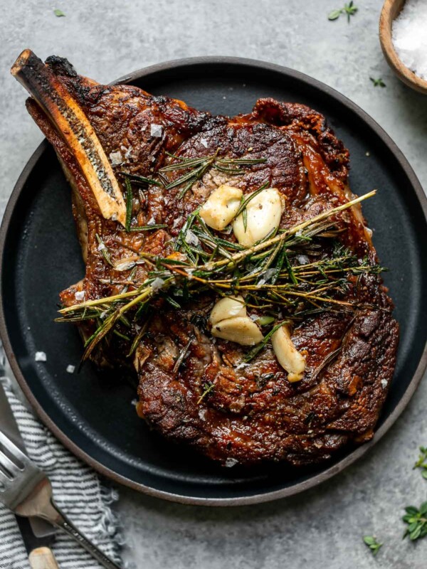 Cooked steak shown on a small black plate. The steak is topped with butter-basted herbs and garlic. The plate is placed atop a light gray surface, next to a bowl of flaky salt, a striped linen napkin and a fork and steak knife.