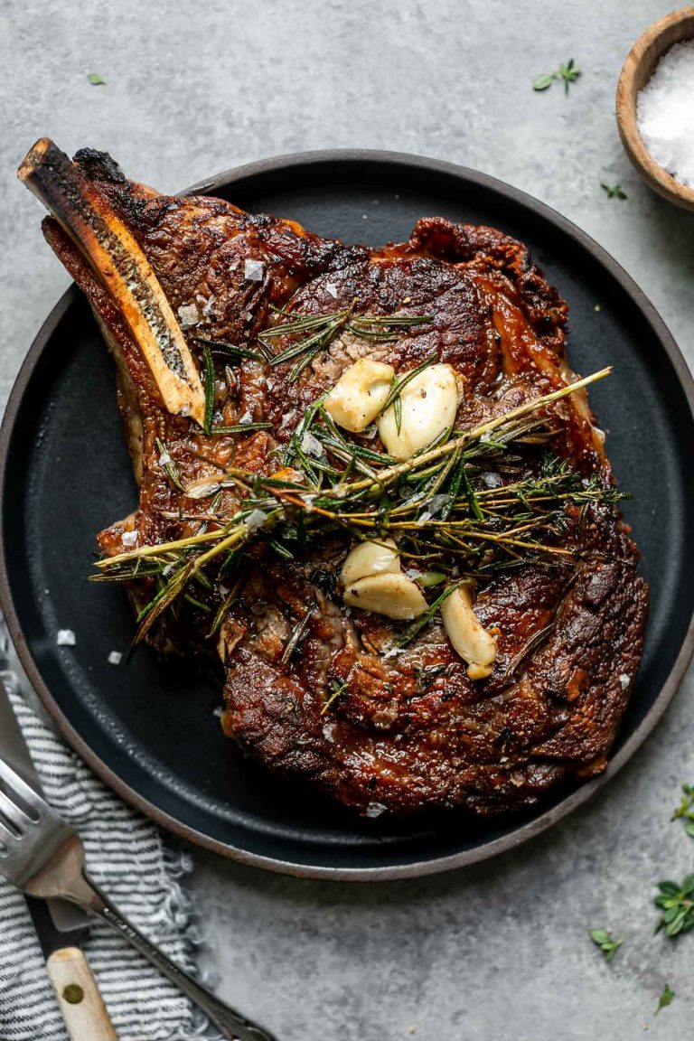 Cooked steak shown on a small black plate. The steak is topped with butter-basted herbs and garlic. The plate is placed atop a light gray surface, next to a bowl of flaky salt, a striped linen napkin and a fork and steak knife.