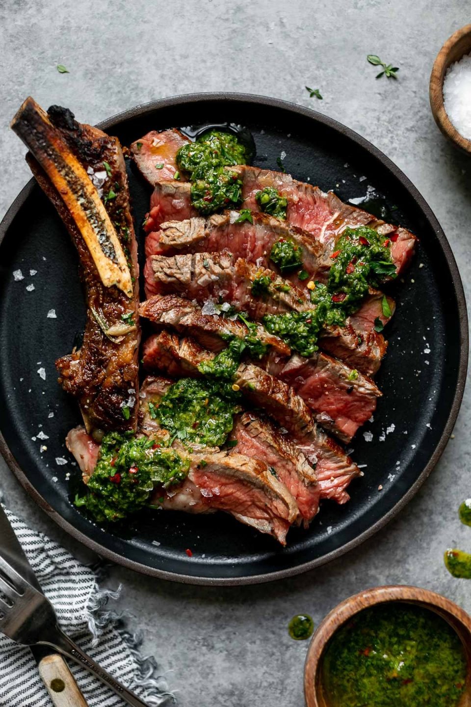 Cooked cast iron steak shown on a small black plate. The steak is sliced off the bone, showing a medium-rare interior, and topped with chimichurri. The plate is placed atop a light gray surface, next to a bowl of flaky salt, a striped linen napkin and a fork and steak knife.
