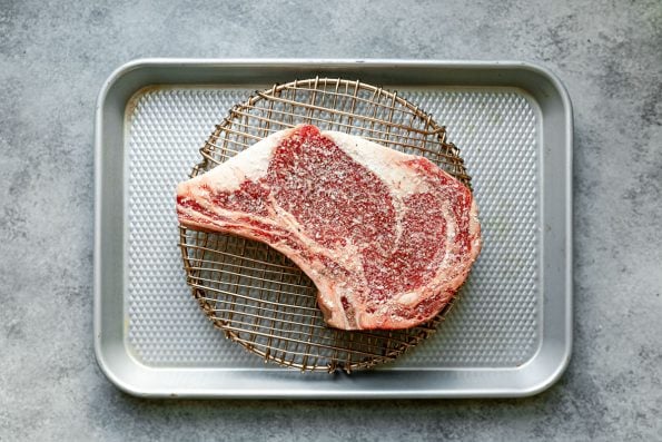An overhead shot of a bone-in ribeye, seasoned generously with salt, placed atop a wire rack on a small baking sheet.