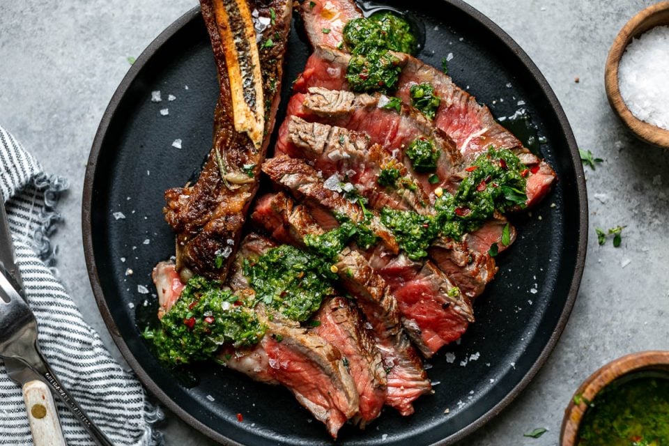Cooked cast iron steak shown on a small black plate. The steak is sliced off the bone, showing a medium-rare interior, & topped with chimichurri. The plate is placed atop a light gray surface, next to a bowl of flaky salt, a striped linen napkin & a fork & steak knife.