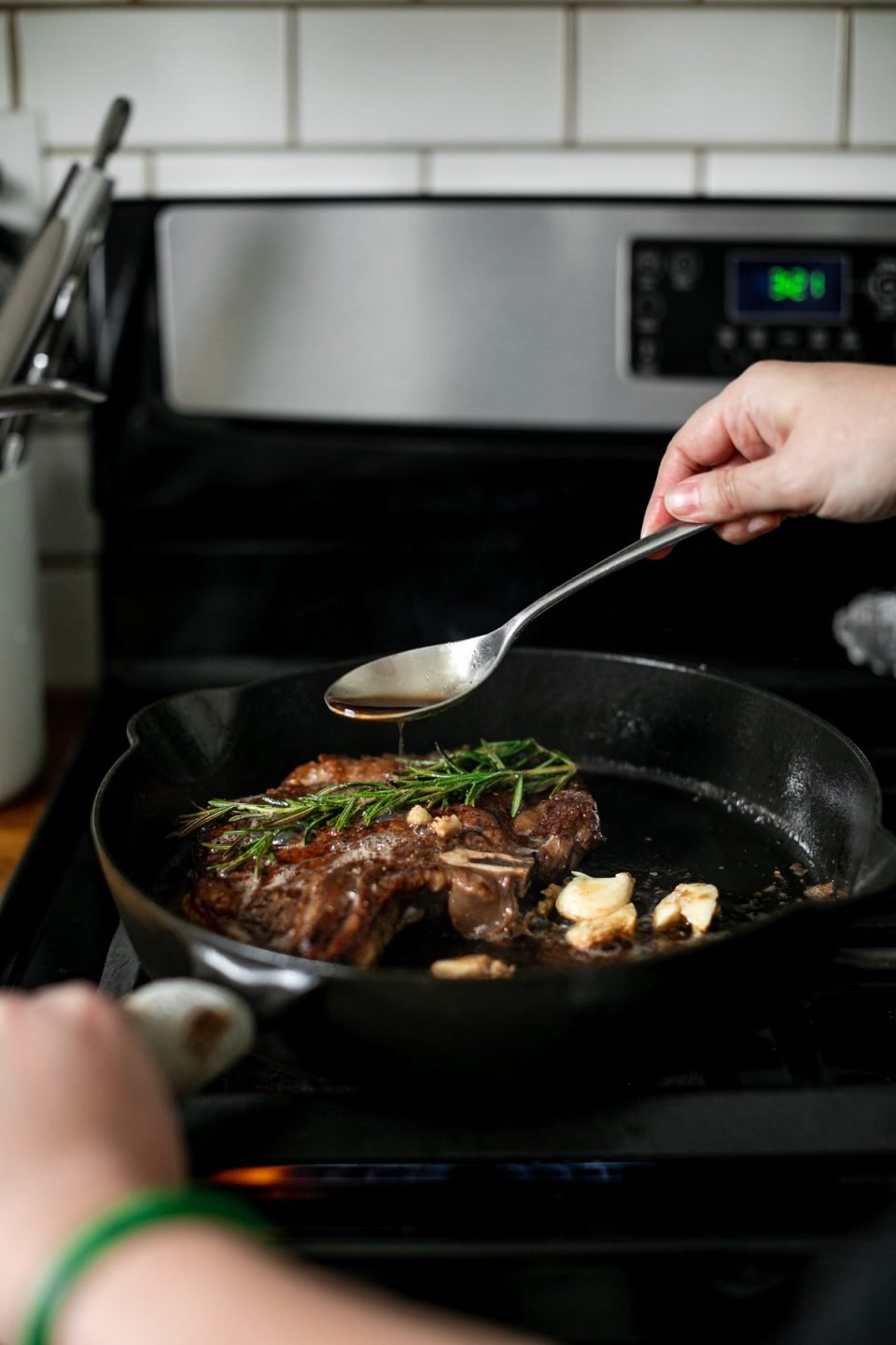 How to cook steak in cast iron skillet: Finishing cast iron steak with butter baste. A woman's hands hold handle of skillet & spoon, spooning browned butter over the steak, which is topped with fresh herbs & crushed garlic cloves.