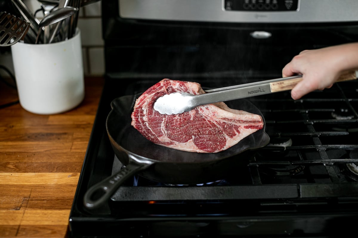 A side angle photo of rendering fat cap off of bone-in ribeye in a large black skillet over stove burner.