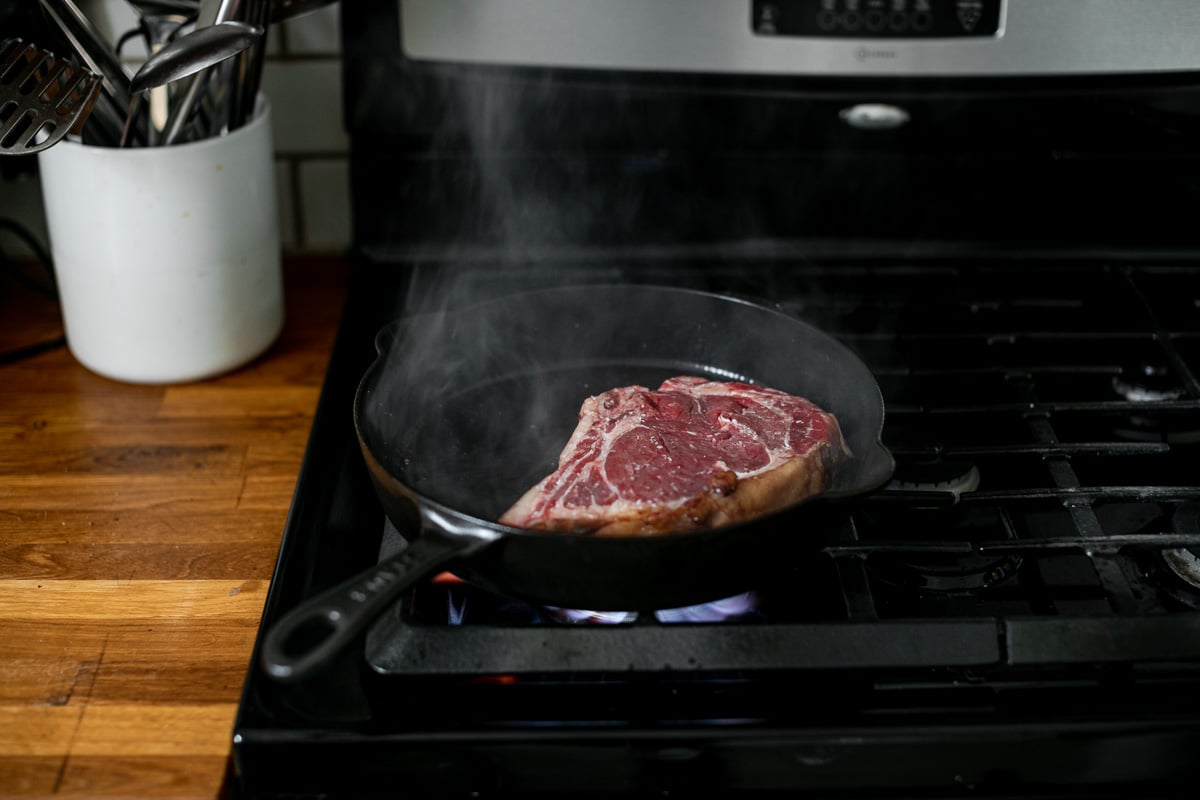 How to cook steak in cast iron skillet: Building crusty sear on bone-in ribeye using frequent turning method in large black cast iron skillet over a stove burner.