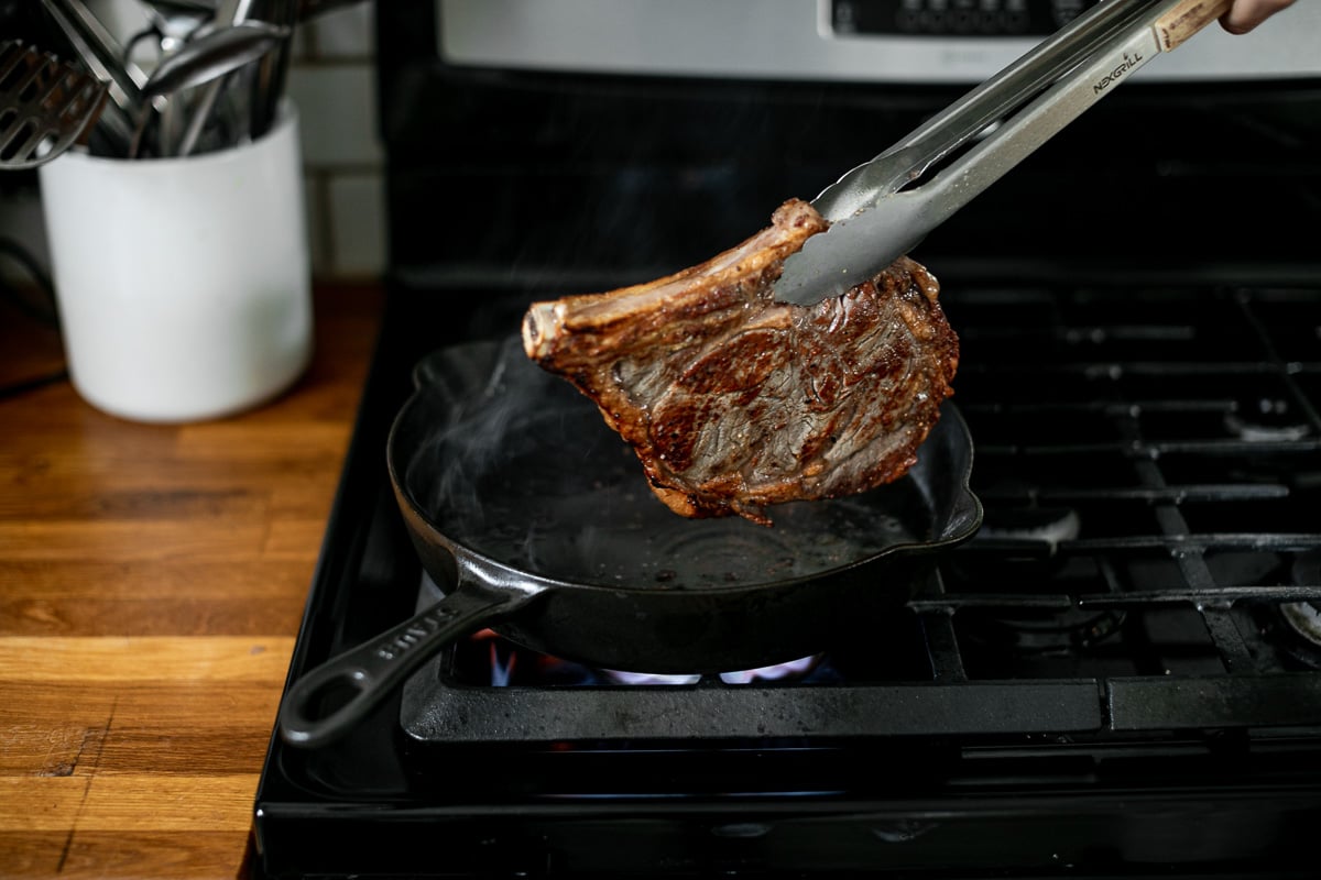 A pair of tongs turns a partially seared steak in a cast iron skillet over high heat on a gas range.
