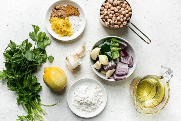 Green falafel ingredients arranged on a white surface: fresh cilantro & parsley, red onion, jalapeno pepper, garlic, flour & baking powder, dried chickpeas, spices, lemon & frying oil.