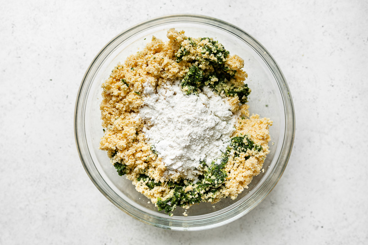 Mixing green falafel - chopped chickpeas, green falafel spice mixture, & flour in a large bowl atop a white surface.