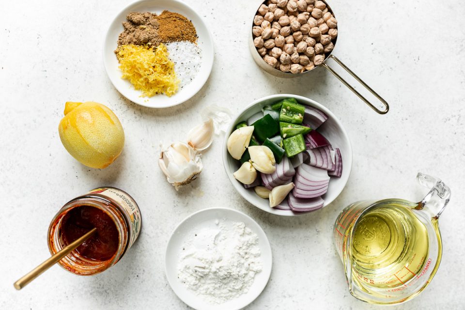 Spicy red falafel ingredients arranged on a white surface: harissa paste, red onion, jalapeno pepper, garlic, flour & baking powder, dried chickpeas, spices, lemon & frying oil.