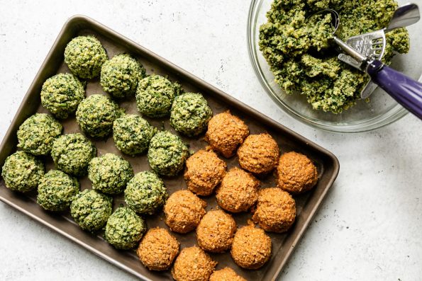 Herby green falafel mixture fills a clear glass mixing bowl. A spring-loaded 2-inch scoop rests inside of the bowl while a baking sheet sits alongside the bowl filled with four rows of four prepared herby green falafel balls and three rows of four spicy red harissa falafel balls. The baking sheet & mixing bowl sit atop a creamy white textured surface.