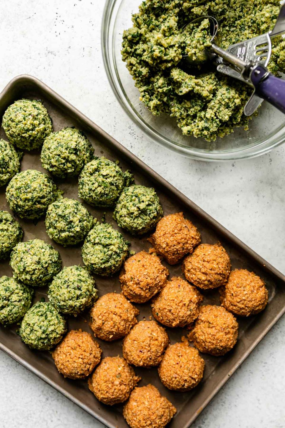 Herby green falafel mixture fills a clear glass mixing bowl. A spring-loaded 2-inch scoop rests inside of the bowl while a baking sheet sits alongside the bowl filled with four rows of four prepared herby green falafel balls and three rows of four spicy red harissa falafel balls. The baking sheet & mixing bowl sit atop a creamy white textured surface.