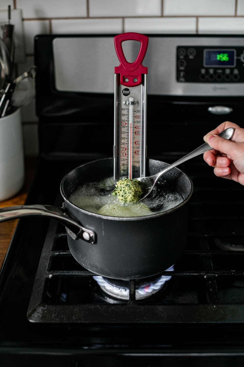 A woman's hand holds a silver spoon with a single uncooked herb green falafel ball resting atop uses the spoon to lower the falafel ball into a black sauce pan filled with prepared oil for frying. A candy thermometer rests inside of the sauce pan on top of a gas stovetop range.