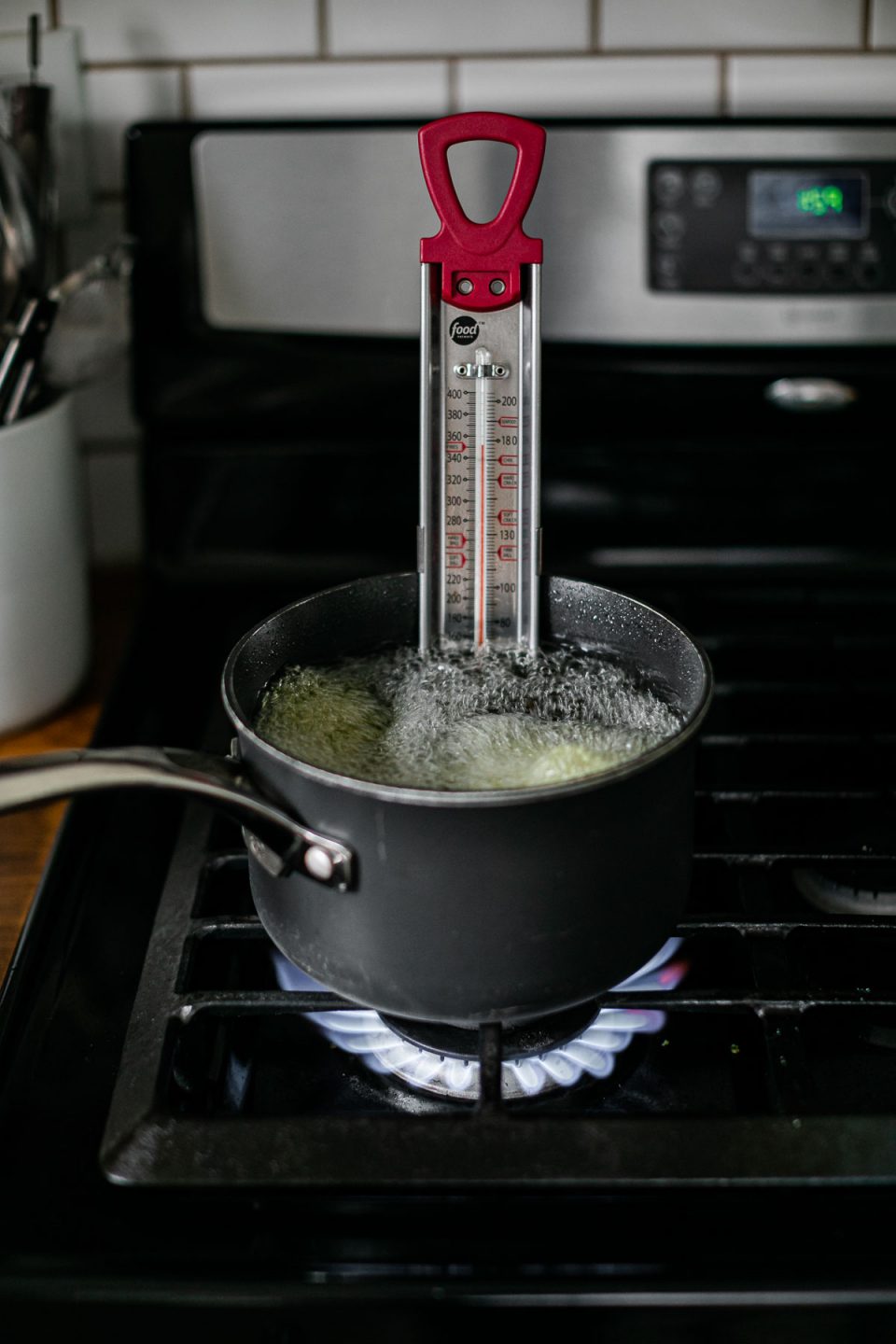 A black sauce pan filled with prepared oil for frying cooks a batch of herb green falafel balls. A candy thermometer rests inside of the sauce pan on top of a gas stovetop range. A crock filled with kitchen utensils sits atop an adjacent butcher block countertop and a white subway tile is shown in the background.