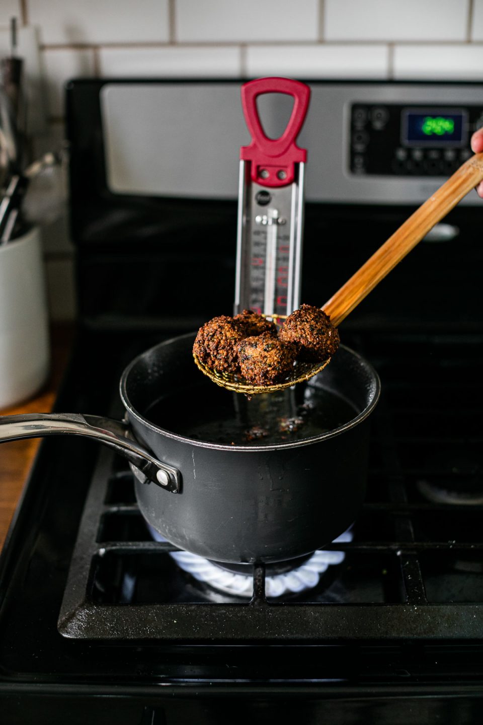 Browned & crispy cooked herb falafel balls are being lifted out of a sauce pan filled with frying oil by a spider strainer being held by a woman's hand. A candy thermometer rests inside of the sauce pan on top of a gas stovetop range. A crock filled with kitchen utensils sits atop an adjacent butcher block countertop and a white subway tile is shown in the background.
