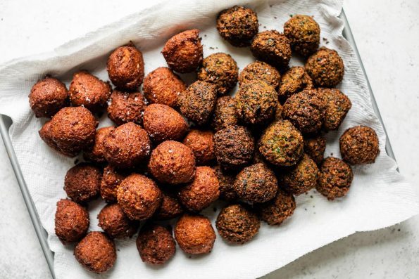 Browned & crispy cooked spicy red harissa and herby green falafel balls arranged on a paper towel-lined baking sheet. The baking sheet sits atop a creamy white textured surface.