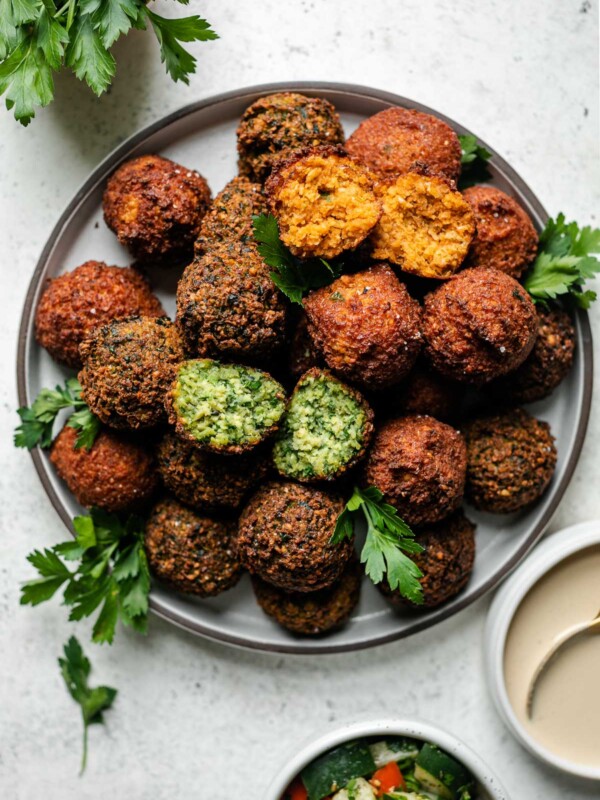 Browned & crispy cooked spicy red harissa and herby green falafel balls arranged on a gray ceramic plate. The plate sits atop a creamy white textured surface and is garnished with fresh parsley leaves. Two small white bowls filled with seasoned diced cucumber and tomatoes and tahini sauce, respectively surround the plate as well as a small bowl filled with a small bunch of fresh parsley.