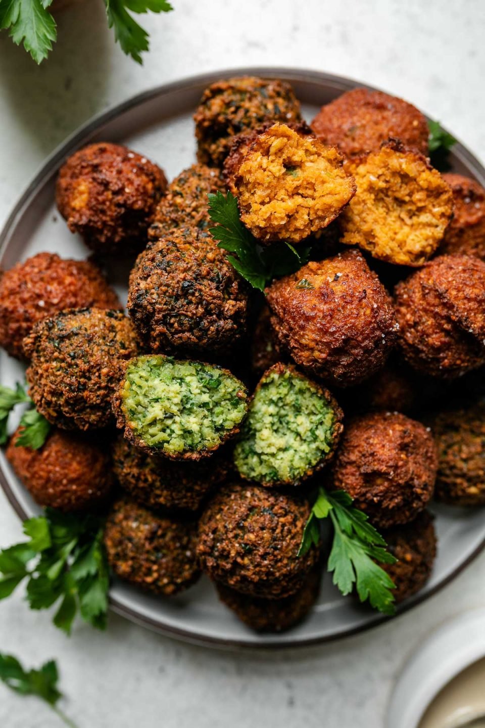 Browned & crispy cooked spicy red harissa and herby green falafel balls arranged on a gray ceramic plate. The plate sits atop a creamy white textured surface and is garnished with fresh parsley leaves. A small white bowl filled with tahini sauce and a small bowl filled with a bunch of fresh parsley surround the plate.