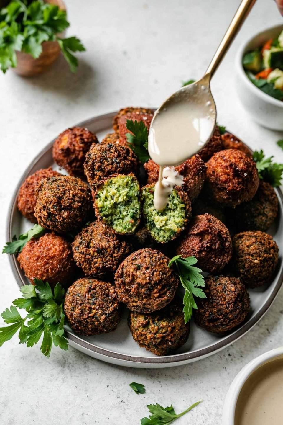 Browned & crispy cooked spicy red harissa and herby green falafel balls arranged on a gray ceramic plate. The plate sits atop a creamy white textured surface and is garnished with fresh parsley leaves. Two small white bowls filled with seasoned diced cucumber and tomatoes and tahini sauce, respectively surround the plate as well as a small bowl filled with a small bunch of fresh parsley. A gold spoon that holds a spoonful of tahini sauce is being held above the plate of falafel & used to drizzle tahini sauce atop the falafel balls.