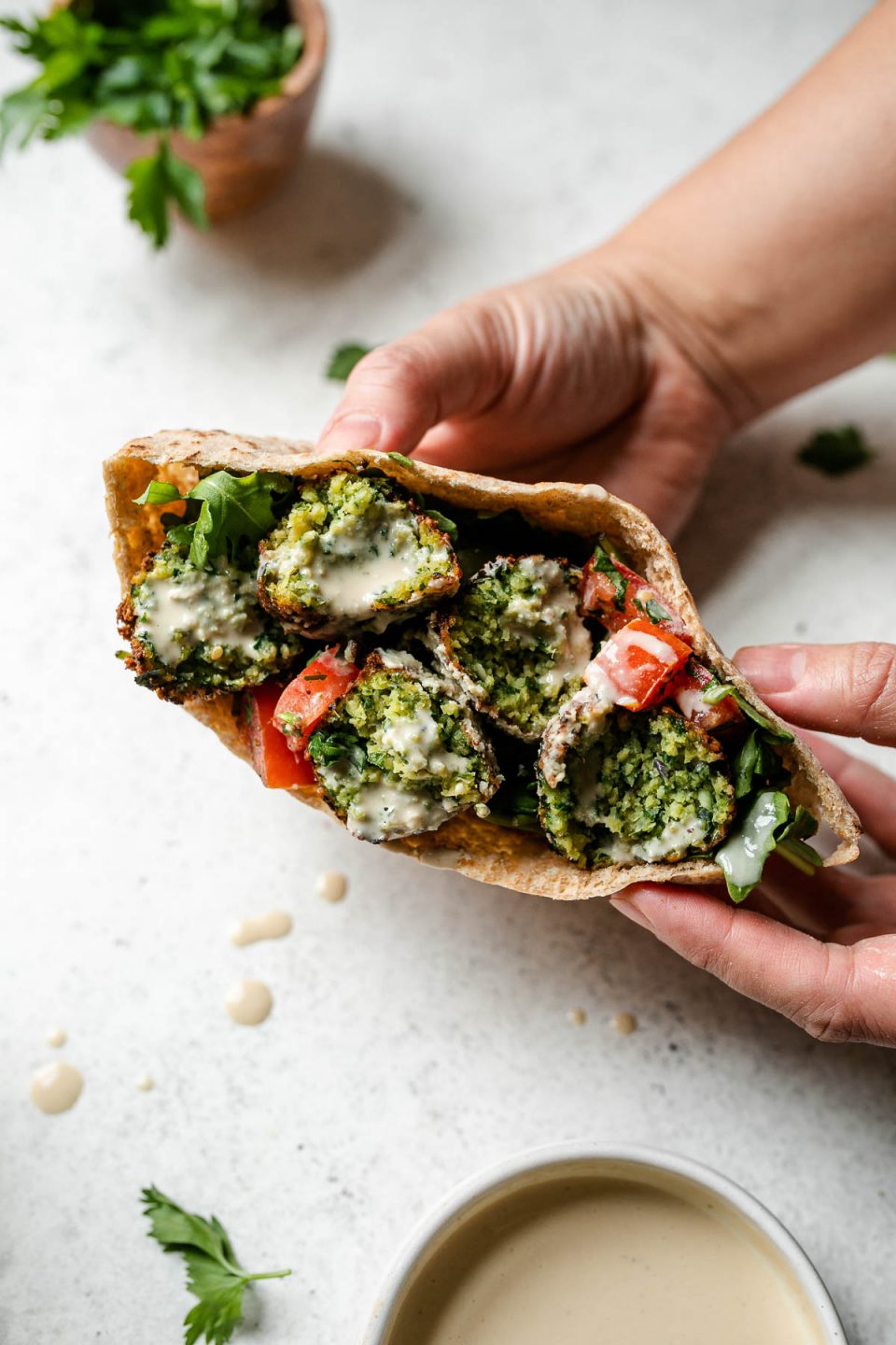 A woman's hands hold a pita filled with crispy herbed green falafel, diced tomatoes, and arugula that has been drizzled with tahini sauce. Her hands rest atop a creamy white textured surface, while loose fresh herbs, a bunch of fresh parsley in a wooden bowl, a small white bowl filled with tahini sauce and drops of spilled tahini sauce surround.