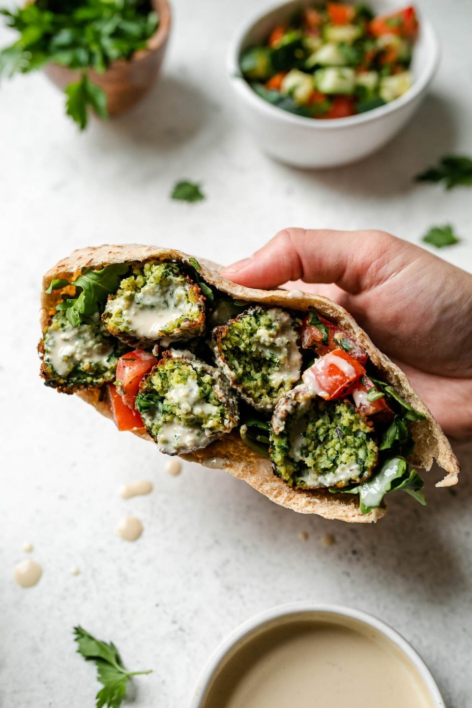 A woman's hands hold a pita filled with crispy herbed green falafel, diced tomatoes, and arugula that has been drizzled with tahini sauce. Her hands rest atop a creamy white textured surface, while loose fresh herbs, a bunch of fresh parsley in a wooden bowl, a small white bowl filled with seasoned cucumbers and tomatoes, a small white bowl filled with tahini sauce and drops of spilled tahini sauce surround.