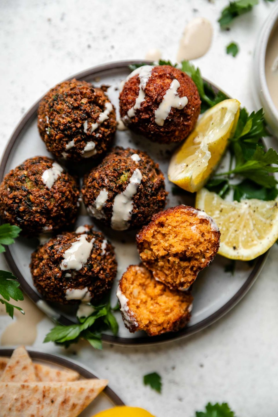 An overhead shot of a small gray ceramic plate filled with homemade crispy falafel balls. The plate is garnished with fresh parsley, lemon wedges, and a drizzle of tahini sauce. A small white bowl filled with tahini sauce and another small gray ceramic plate filled with pita rests alongside the falafel balls on the plate at center. All items sit atop a creamy white textured surface.