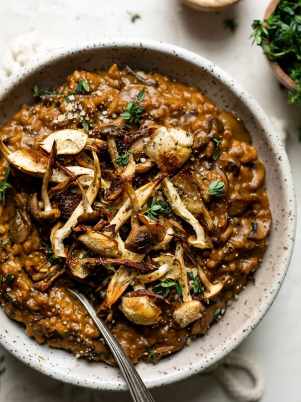 Porcini mushroom risotto shown in a speckled ceramic bowl, topped with crispy roasted mushrooms & fresh thyme leaves. The bowl sits atop a creamy cement surface, next to smaller wooden bowls filled with extra grated parmesan & fresh thyme.