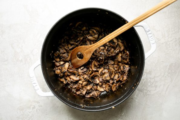 How to make porcini mushroom risotto: Softening sliced cremini mushrooms & shallot in a large white Dutch oven, which sits atop a creamy cement surface.